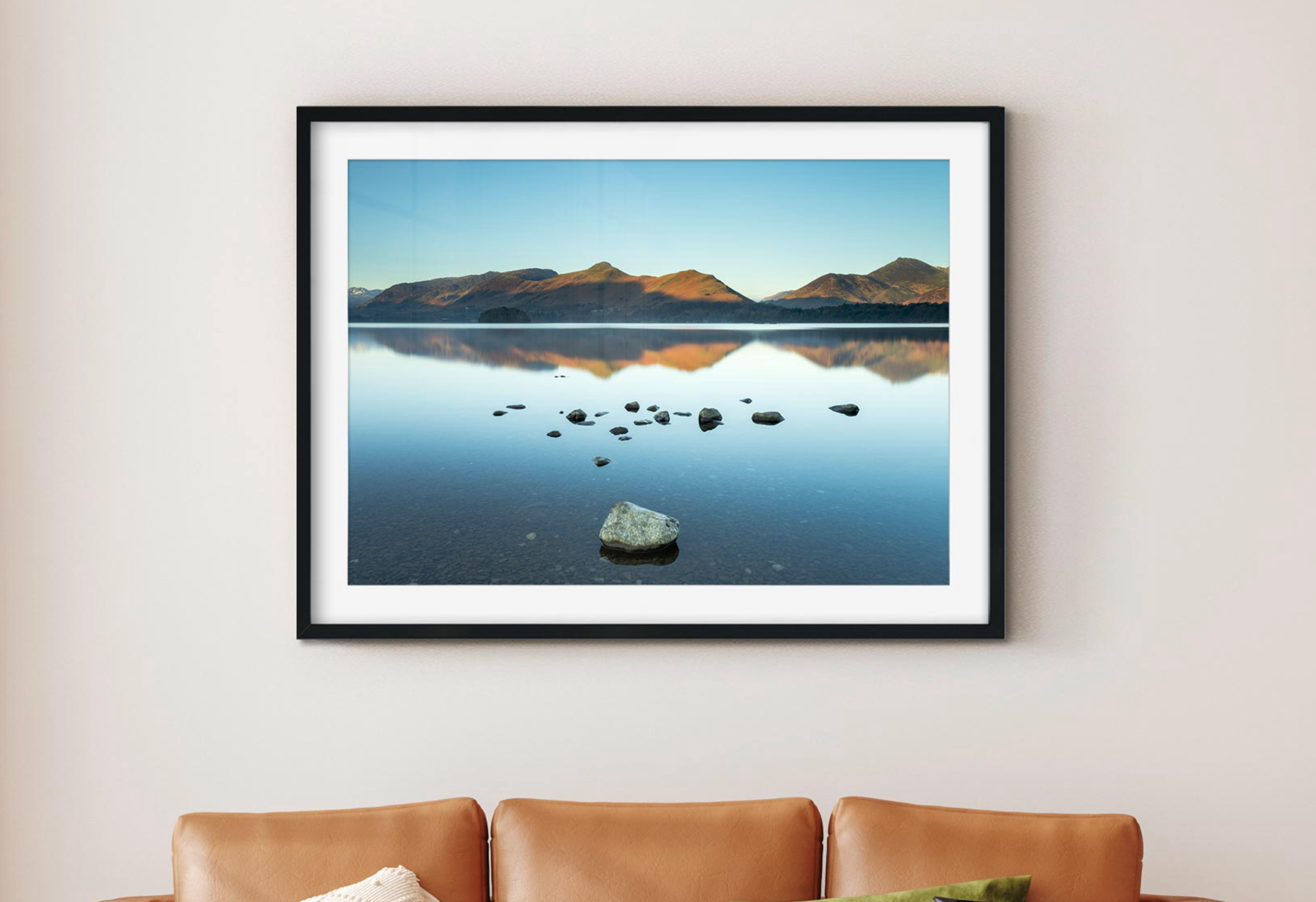 a photo of a reflective lake in a black frame above a tan leather sofa.